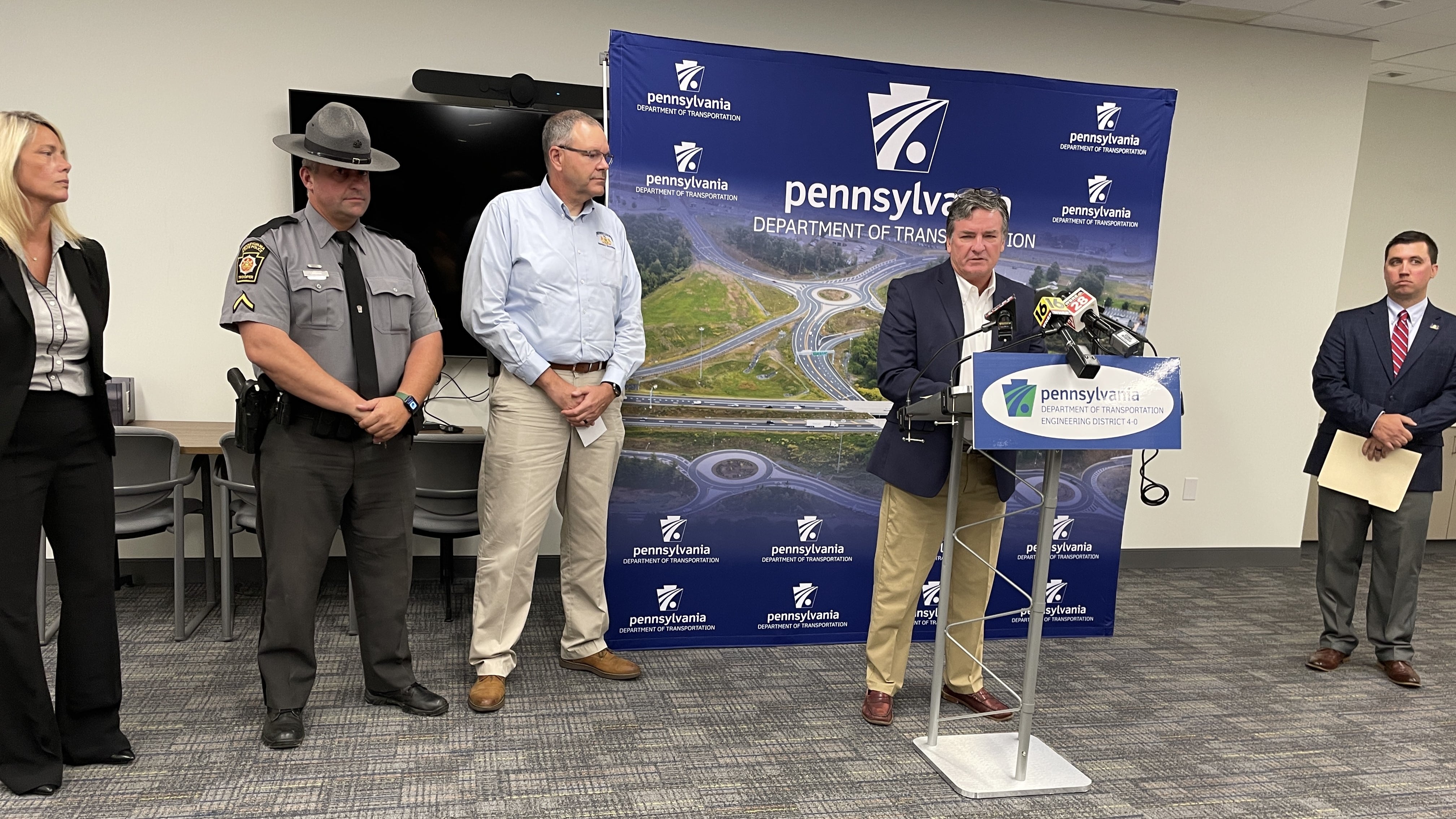 An image of PennDOT Secretary Michael Carroll, joined by other officials, speaking at a press conference from behind a podium with a backdrop behind him showing the PennDOT logo and the words Pennsylvania Department of Transportation